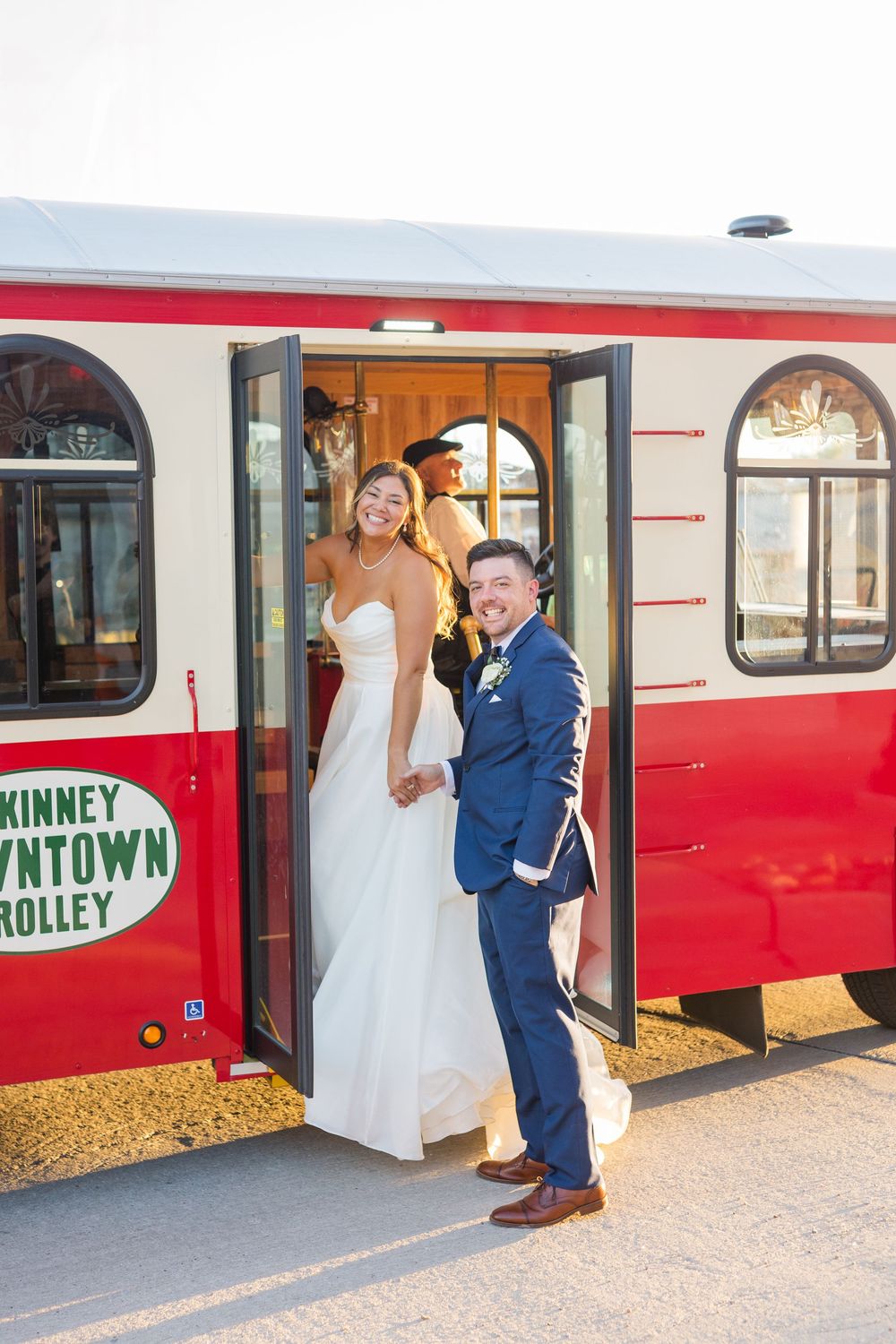 Smiling bride and groom pose at entrance of red McKinney Downtown Trolley on wedding day.