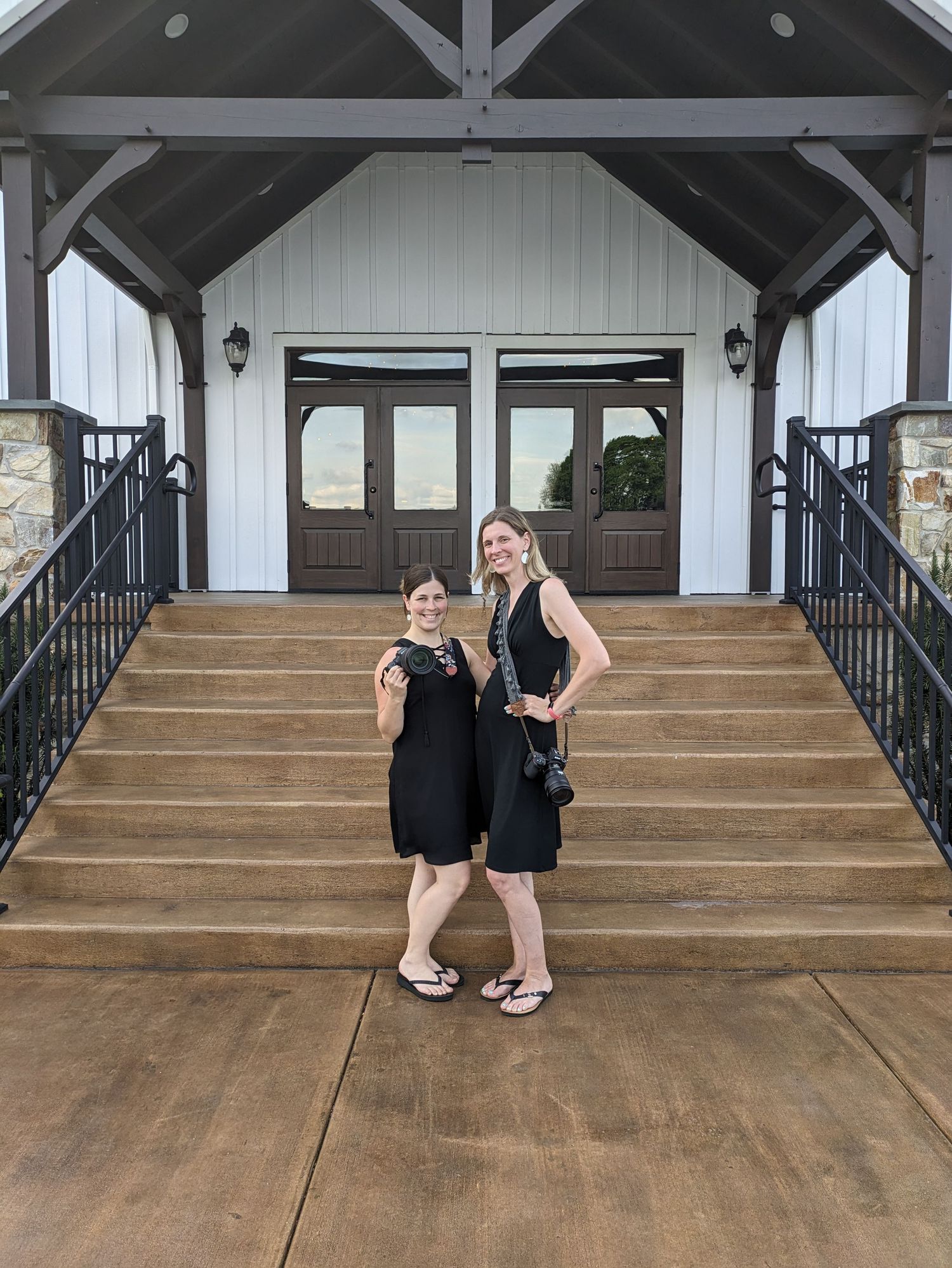Two people in black dresses pose together on concrete steps leading to a white building entrance with double doors.