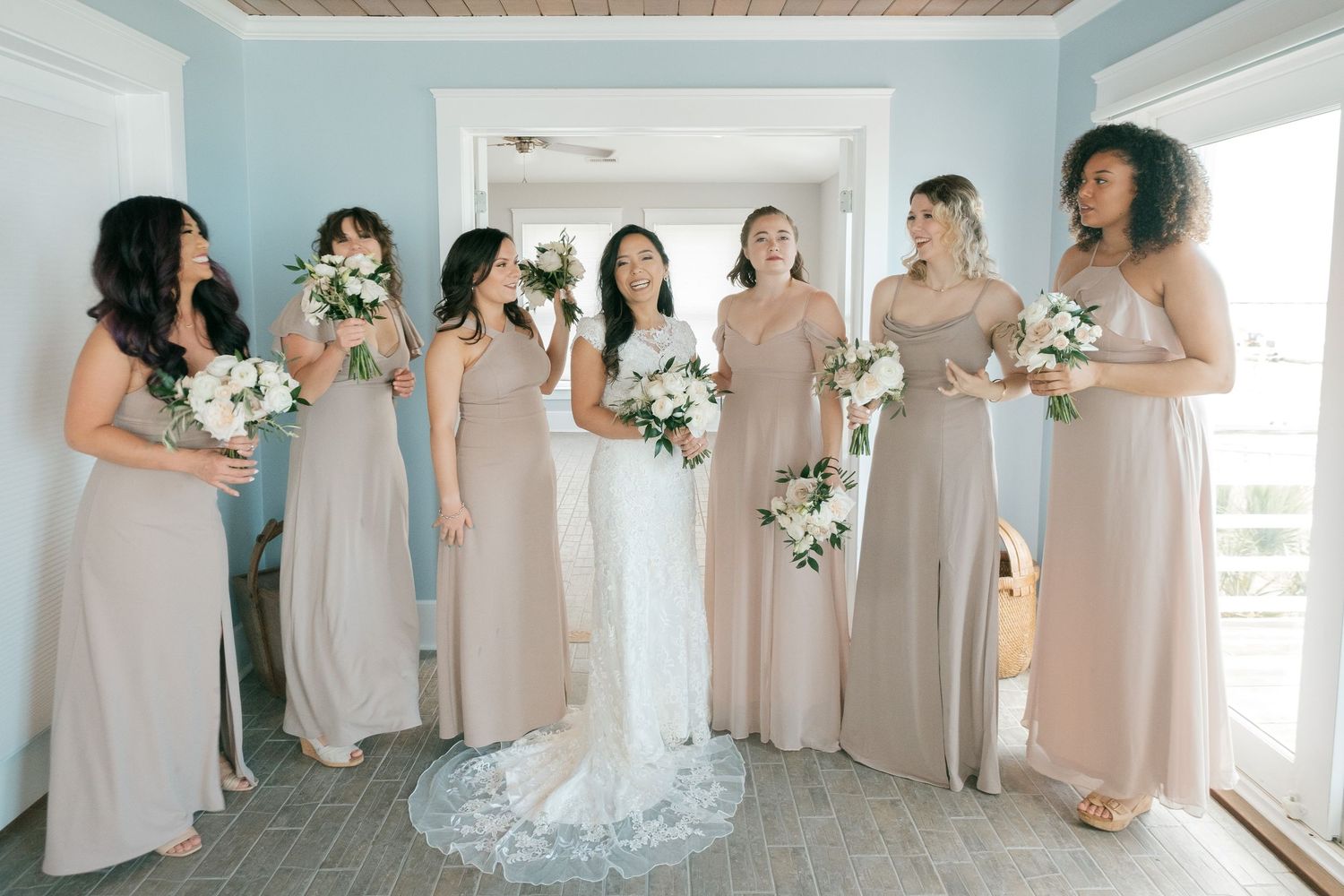 A bridal party wearing elegant floor-length beige dresses poses together holding white floral bouquets in a bright room.