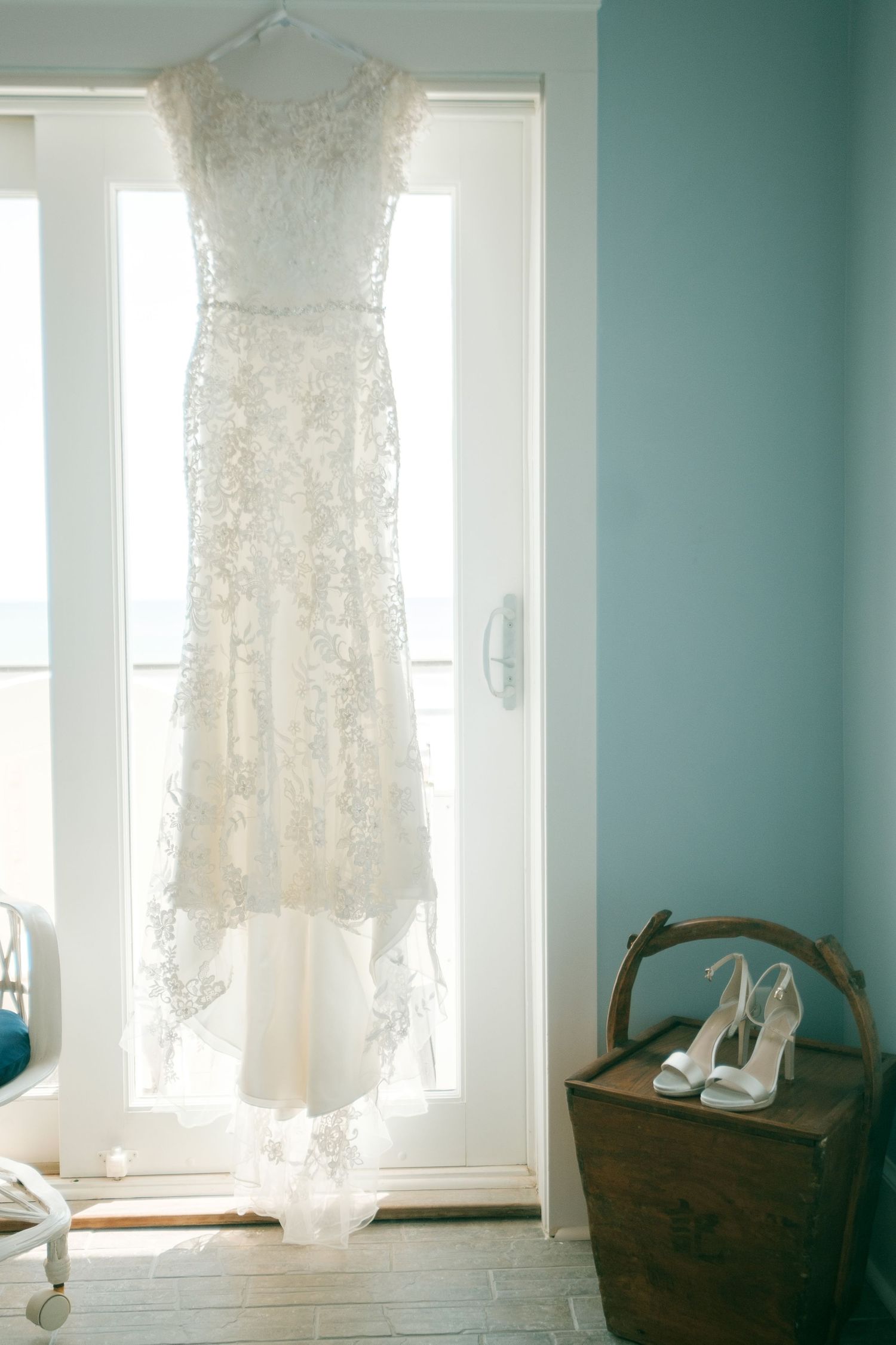 A delicate white lace wedding dress hangs in front of a window with white heels on a vintage wooden chest.