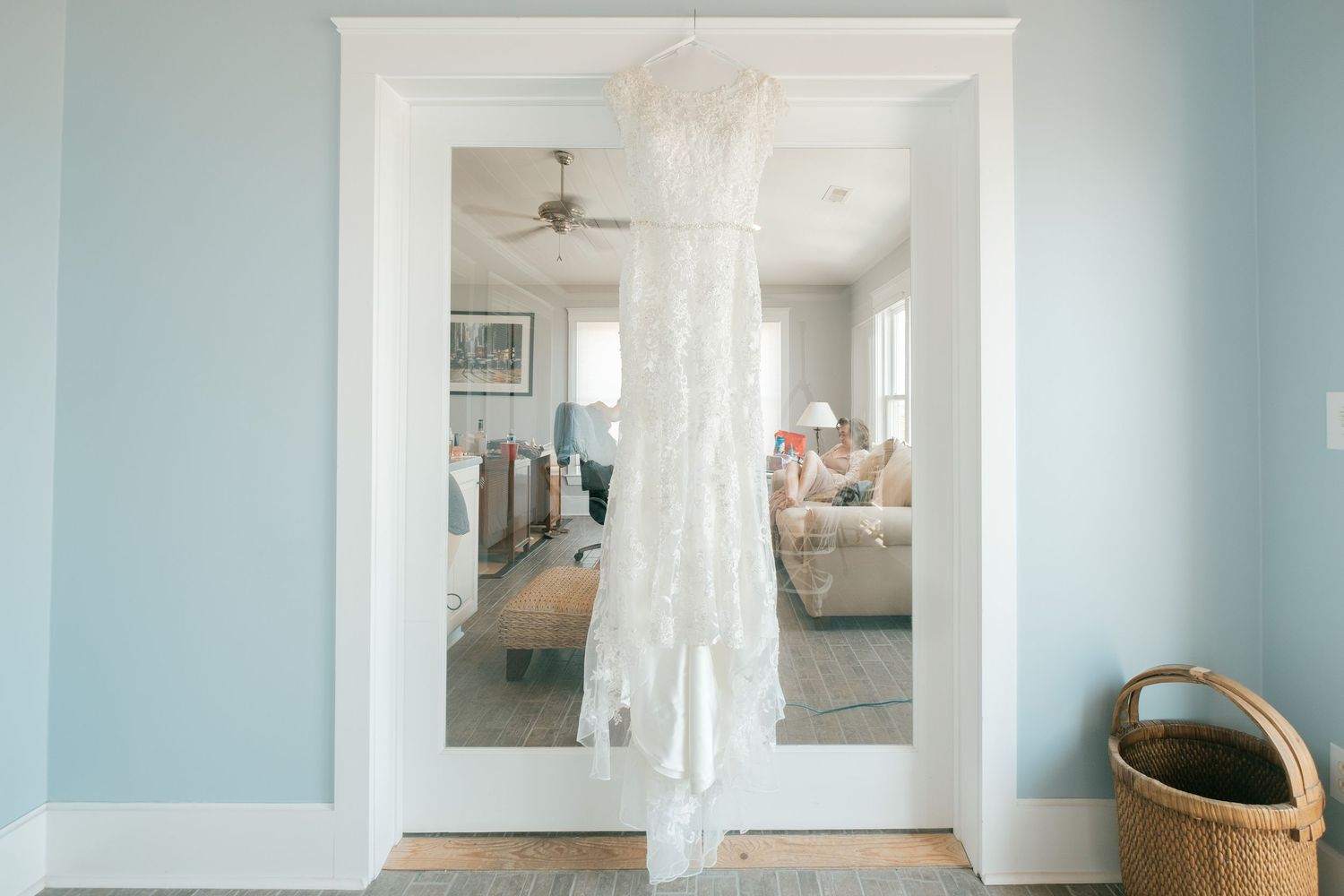 White wedding dress hanging in a doorway of a light blue room with hardwood floors and a ceiling fan.