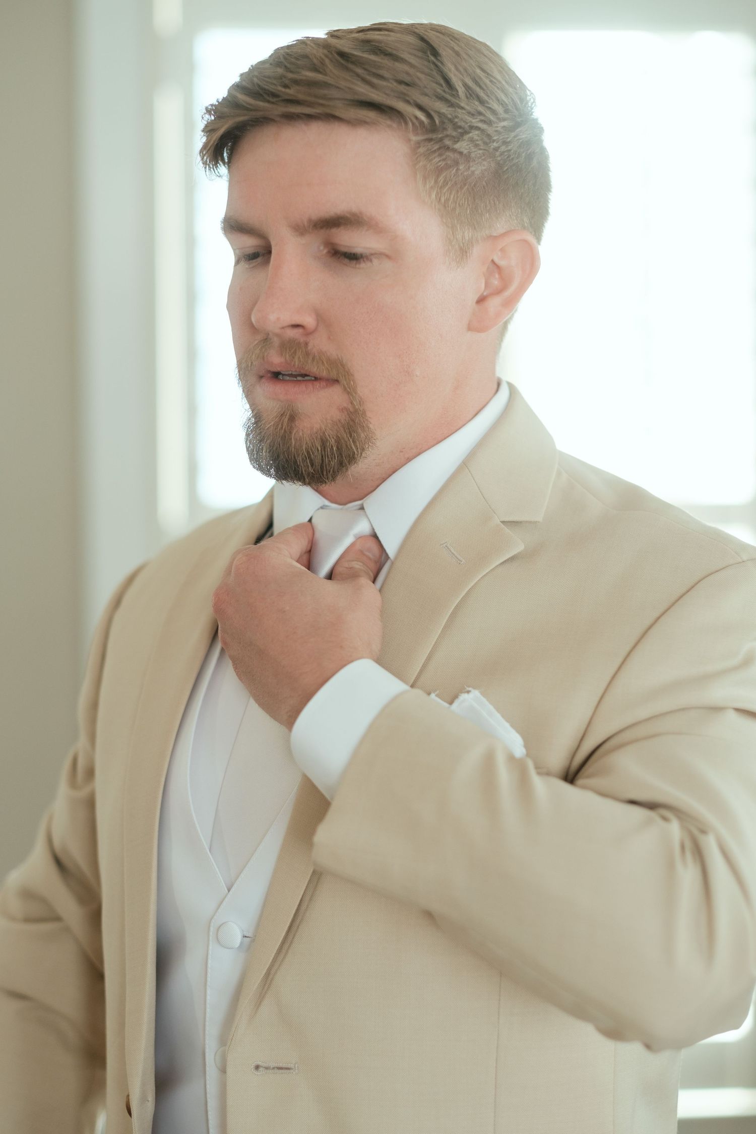 A groom in a beige suit adjusts his tie while getting ready.