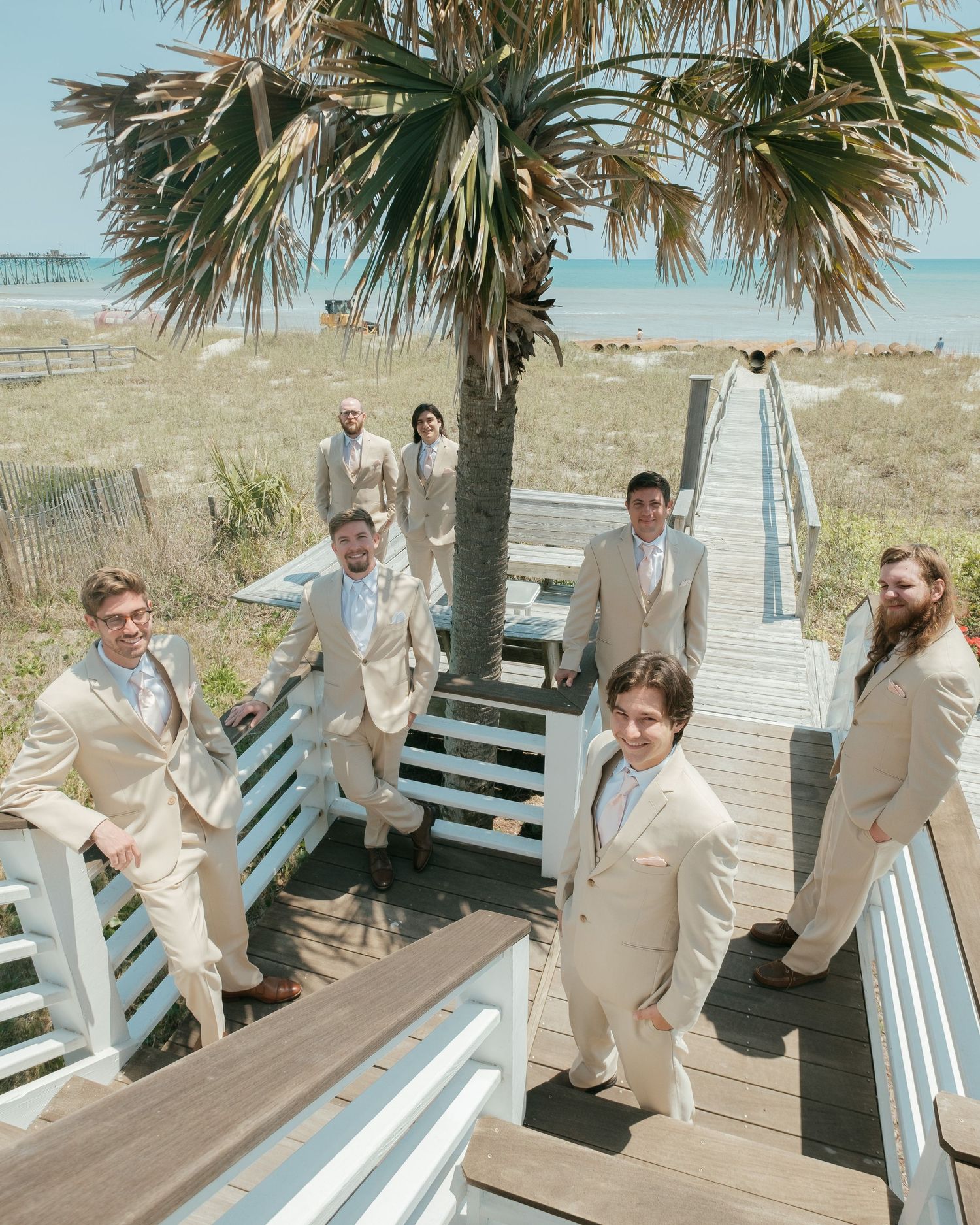 Wedding party poses on a coastal boardwalk with palm trees and ocean in background.
