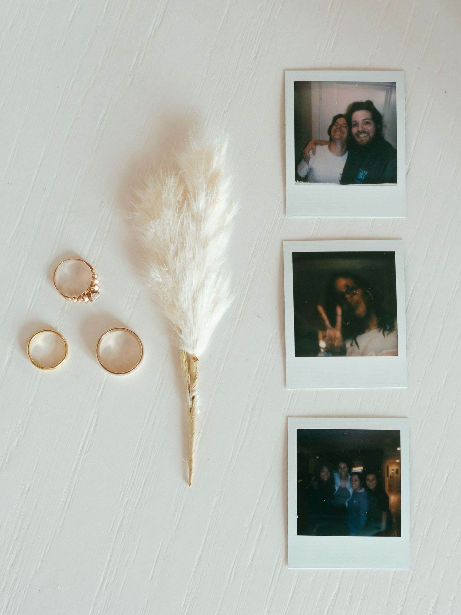 Wedding rings and dried pampas grass displayed alongside vintage polaroid photos on white wood surface.