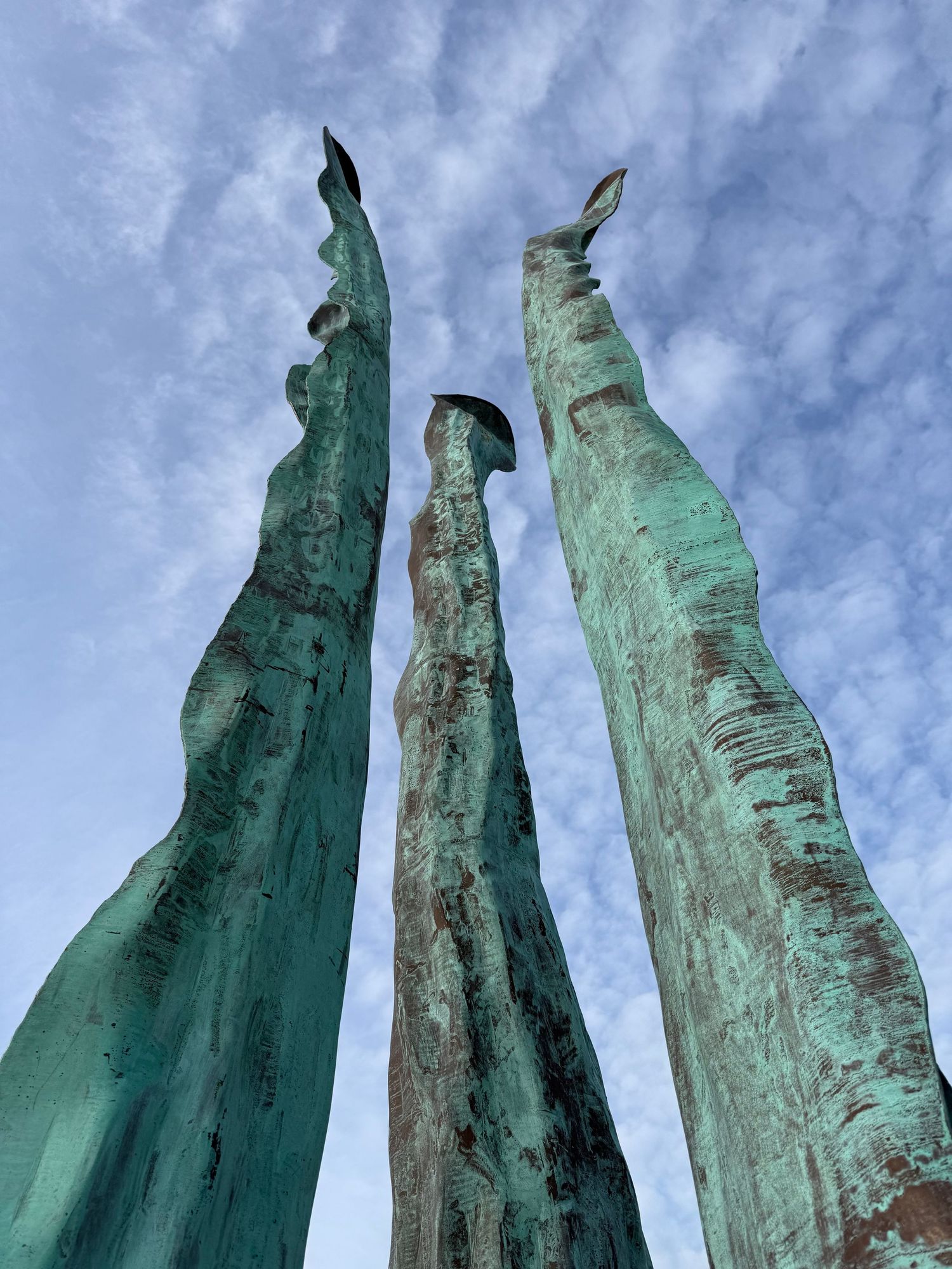 Three tall oxidized copper sculptures with green patina reaching toward a cloudy blue sky.