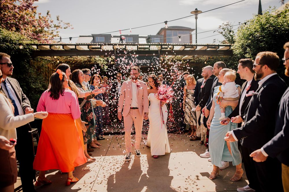 Wedding guests gather outdoors for a sunny celebration on a patio decorated with string lights and floral arrangements.
