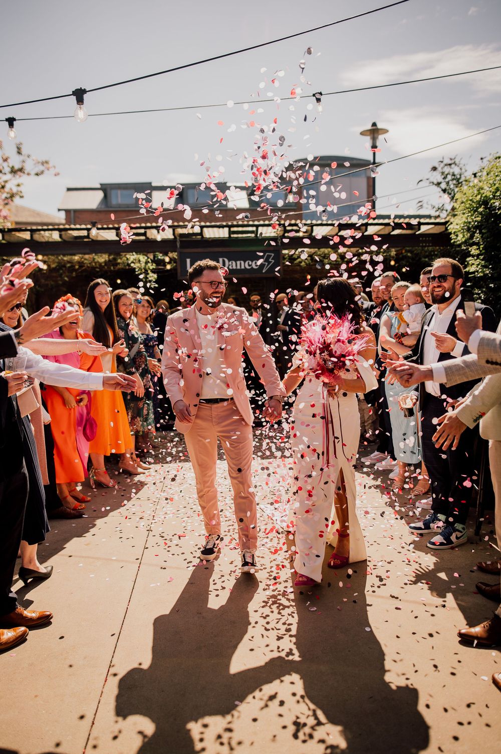 Wedding guests shower newlyweds with pink flower petals during outdoor celebration in warm sunlight.
