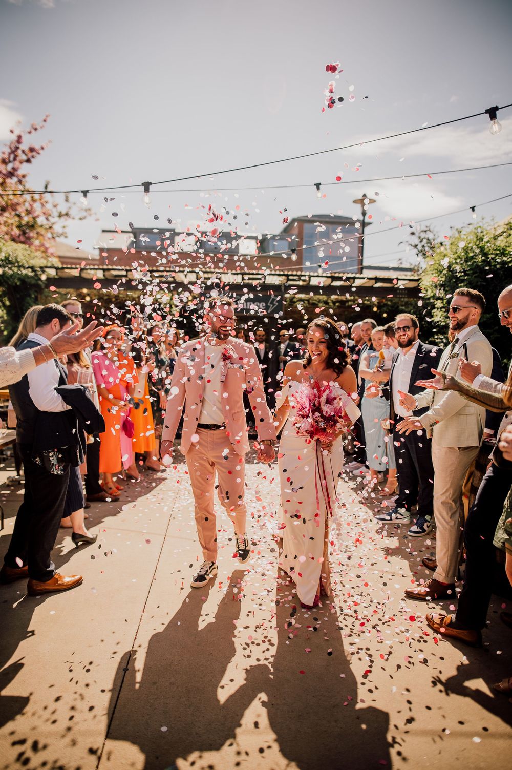 Wedding guests toss pink confetti at a rooftop celebration as string lights hang overhead during a sunset ceremony.