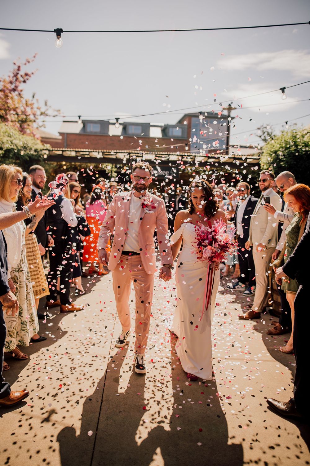 Wedding guests celebrate on an outdoor patio as pink and white confetti rains down during a joyous celebration.