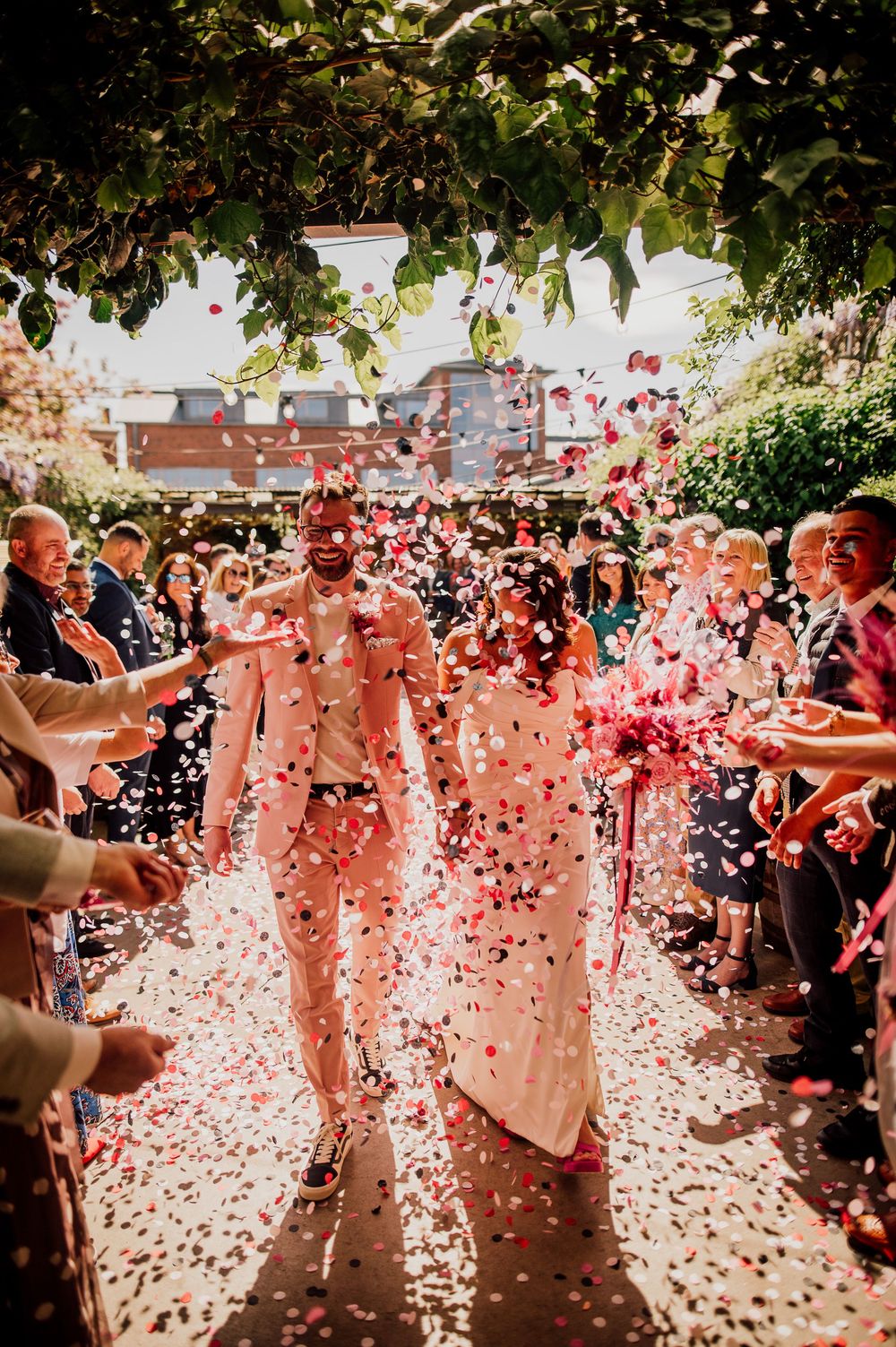 Wedding guests celebrate with pink confetti being thrown in the air during an outdoor celebration on a sunny day.