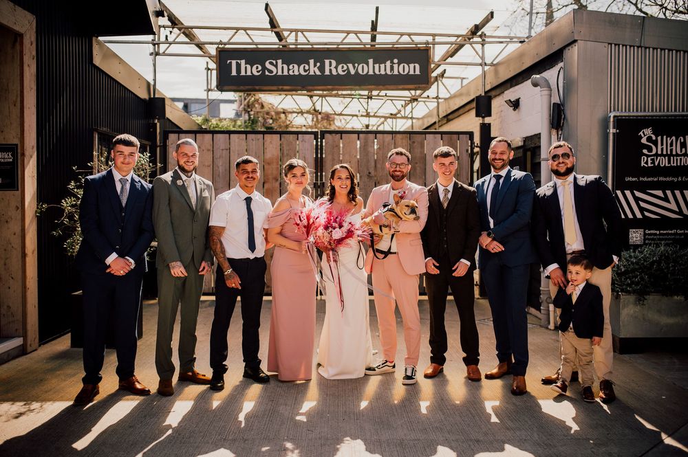 Wedding party members pose together in formal attire at an outdoor venue with rustic wooden beams and decor.