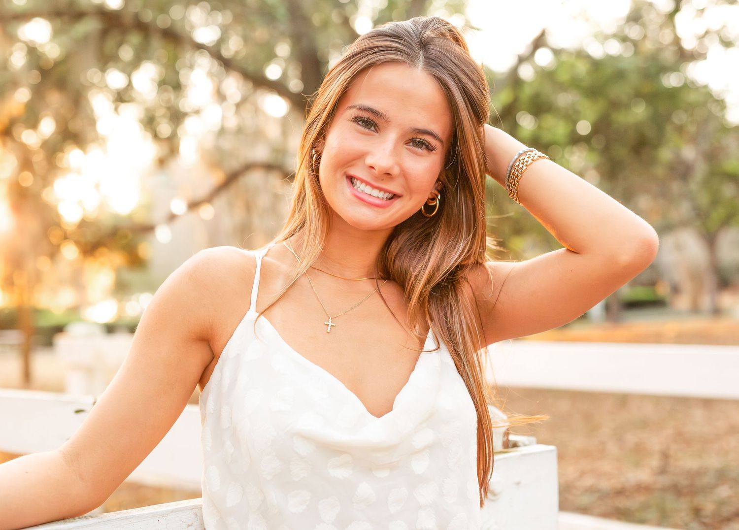 Young woman in white dress poses outdoors during golden hour sunset in a natural setting.