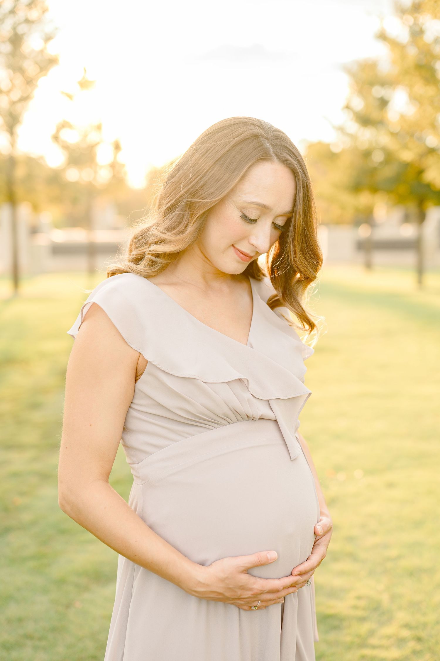A pregnant woman in a flowing beige dress touches her belly while standing in a sunlit park at golden hour houston maternity photographer
