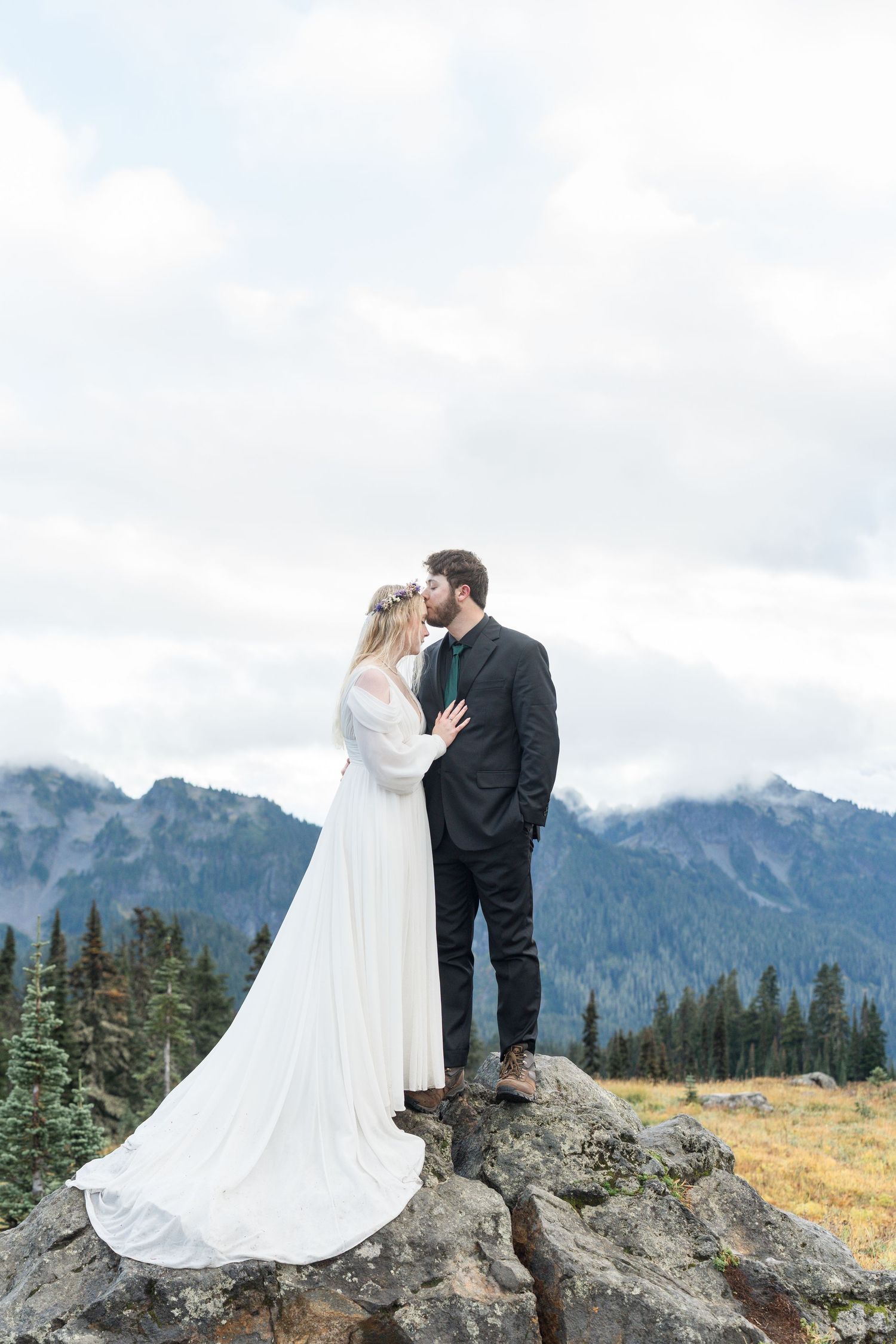 A romantic wedding couple embraces on a mountain peak with dramatic mountain ranges and evergreen forests in the background.