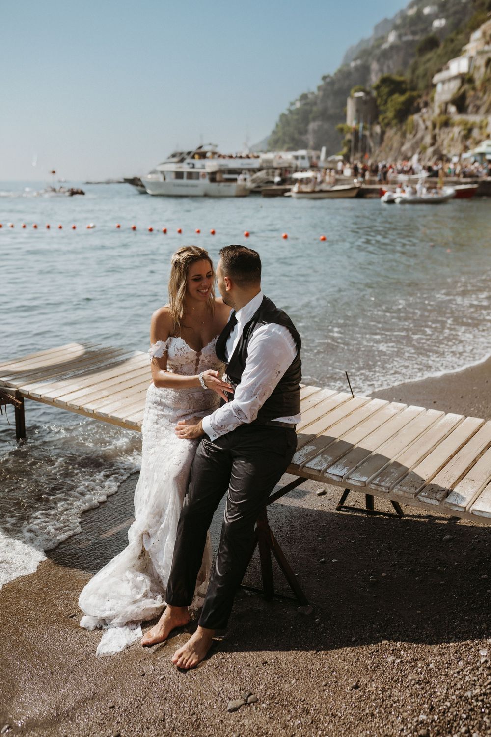 A romantic couple sits together on a wooden dock by the Mediterranean Sea on a sunny coastal afternoon.