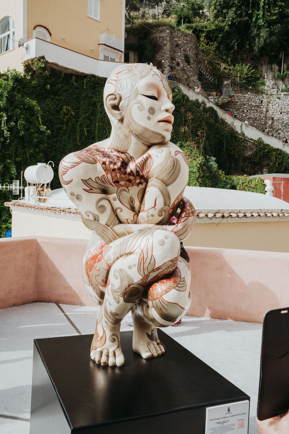 A modern sculptural art piece featuring a crouching figure with intricate body art designs displayed on a black pedestal.