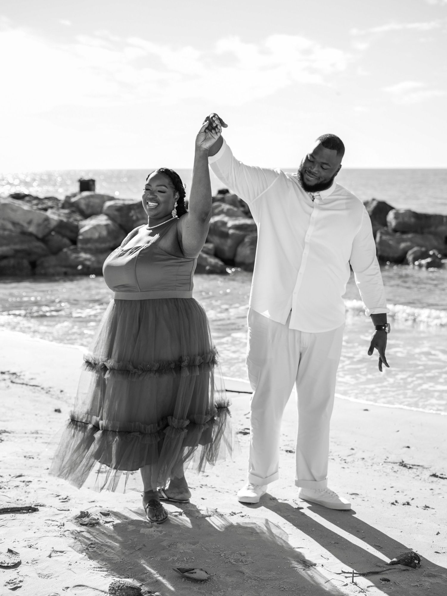 A couple in white clothing and a flowing tulle dress pose together on a sunny beach during a photo session.