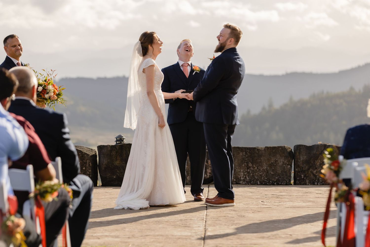 A wedding ceremony takes place on a scenic mountain overlook with mountains and forests in the background.