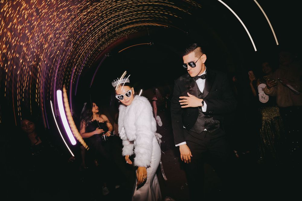Bride in white fur coat and groom in tuxedo walk through illuminated neon tunnel at festive wedding reception.
