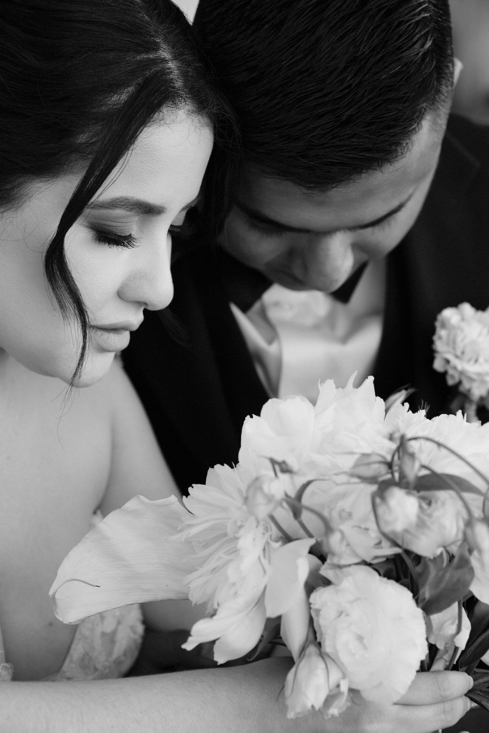 Black and white wedding photo of bride and groom leaning together over a bouquet of peonies.