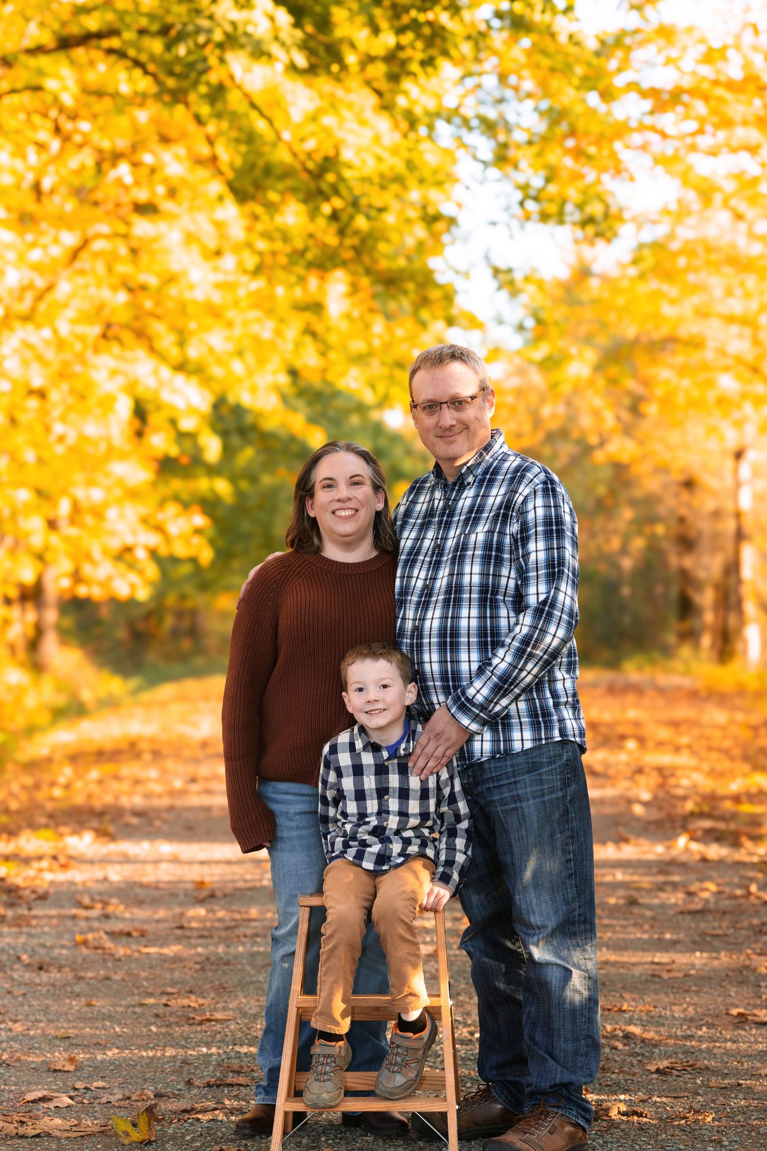 Family of three posing on a tree-lined path surrounded by vibrant yellow autumn foliage during fall season.
