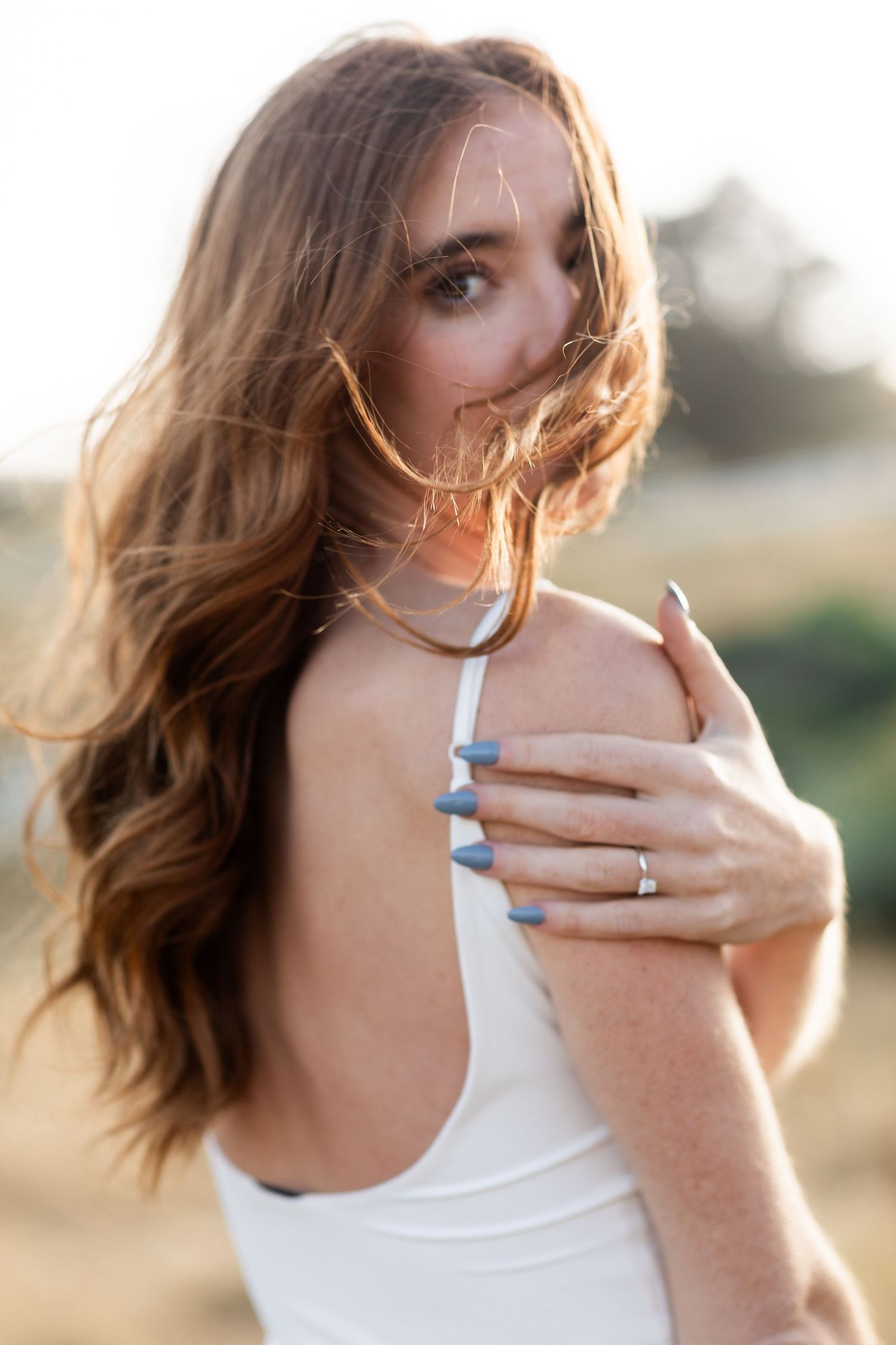 A woman in a white tank top poses in soft golden sunlight with windswept hair and a ring on her finger.