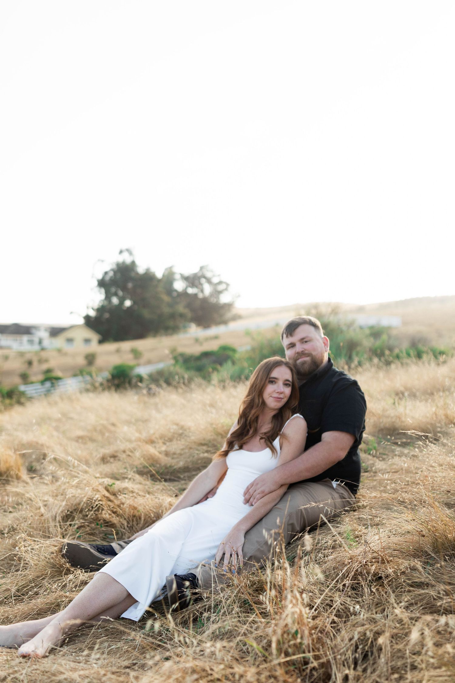 A couple relaxes together on a grassy hillside during a sunny afternoon photoshoot.
