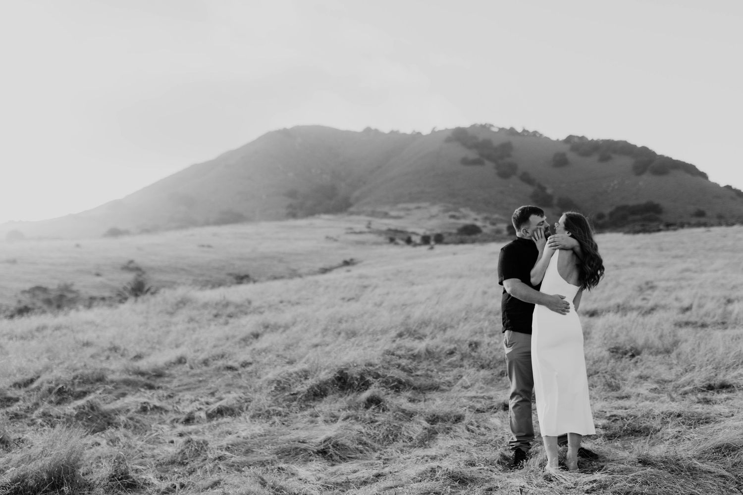A romantic couple embraces on a hillside in a black and white photograph with rolling hills in the background.