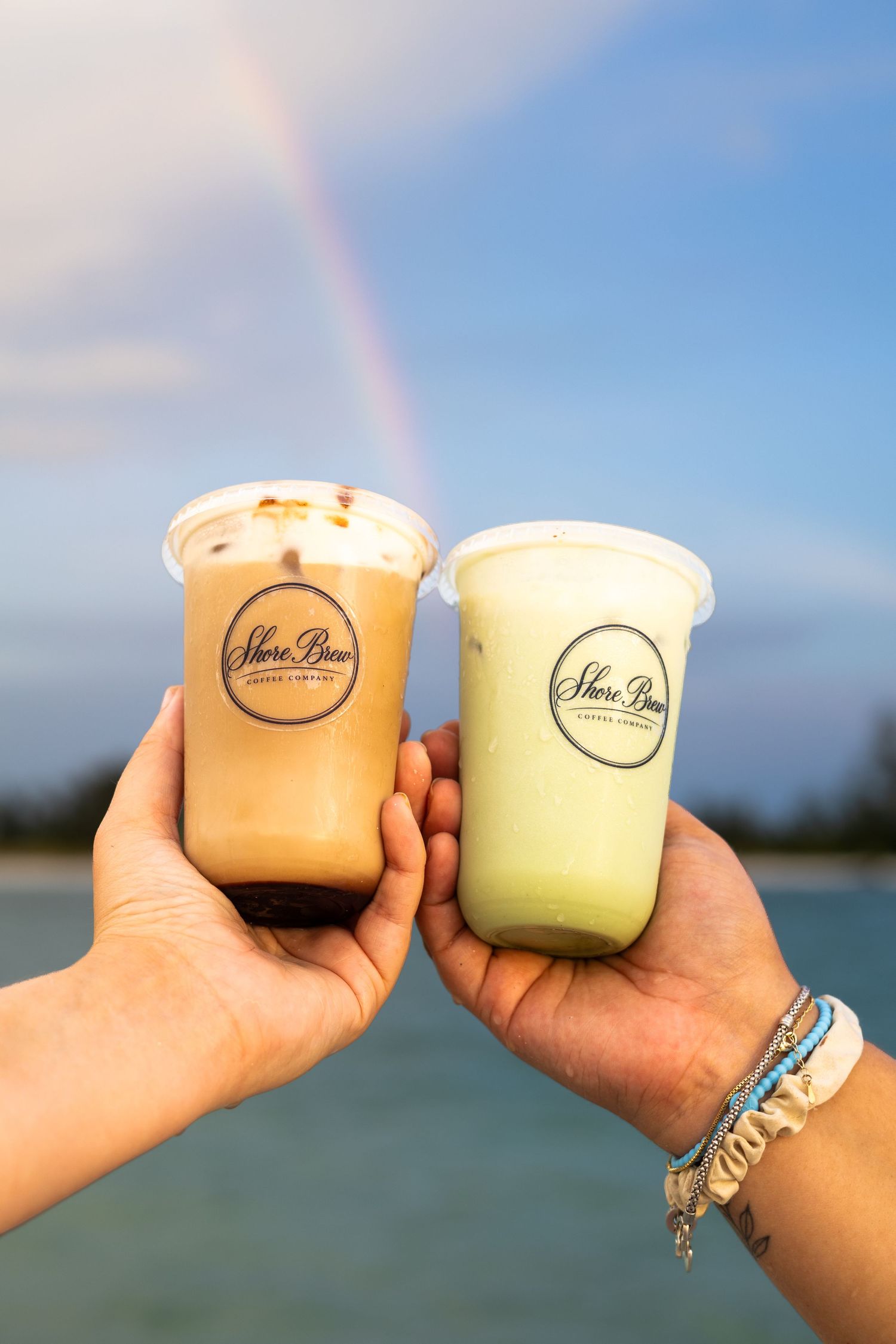 Two hands holding Shore Brew iced coffee drinks with a rainbow and ocean in the background.