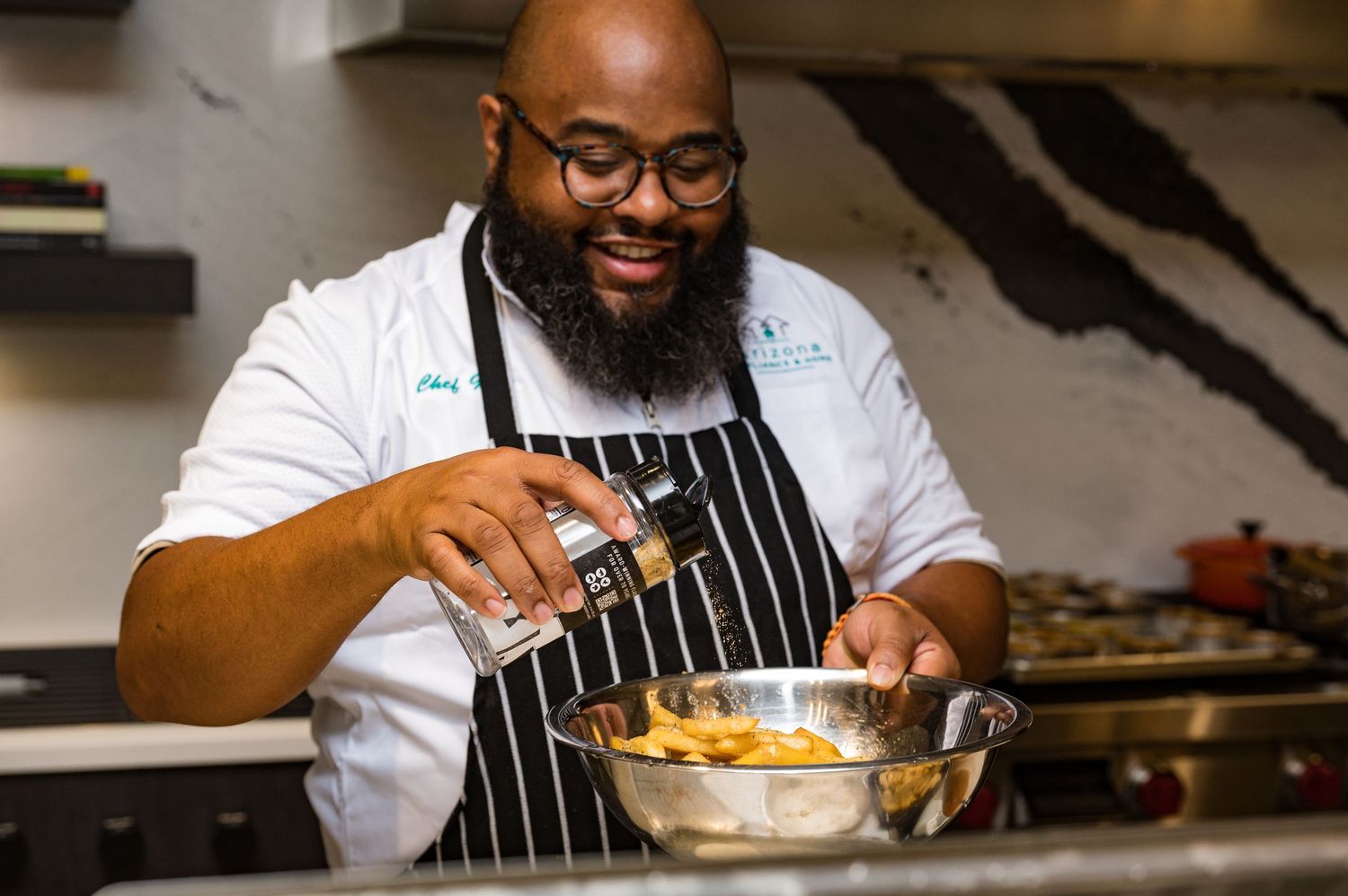 Smiling chef in white coat and striped apron seasoning food in a steel mixing bowl in a professional kitchen.