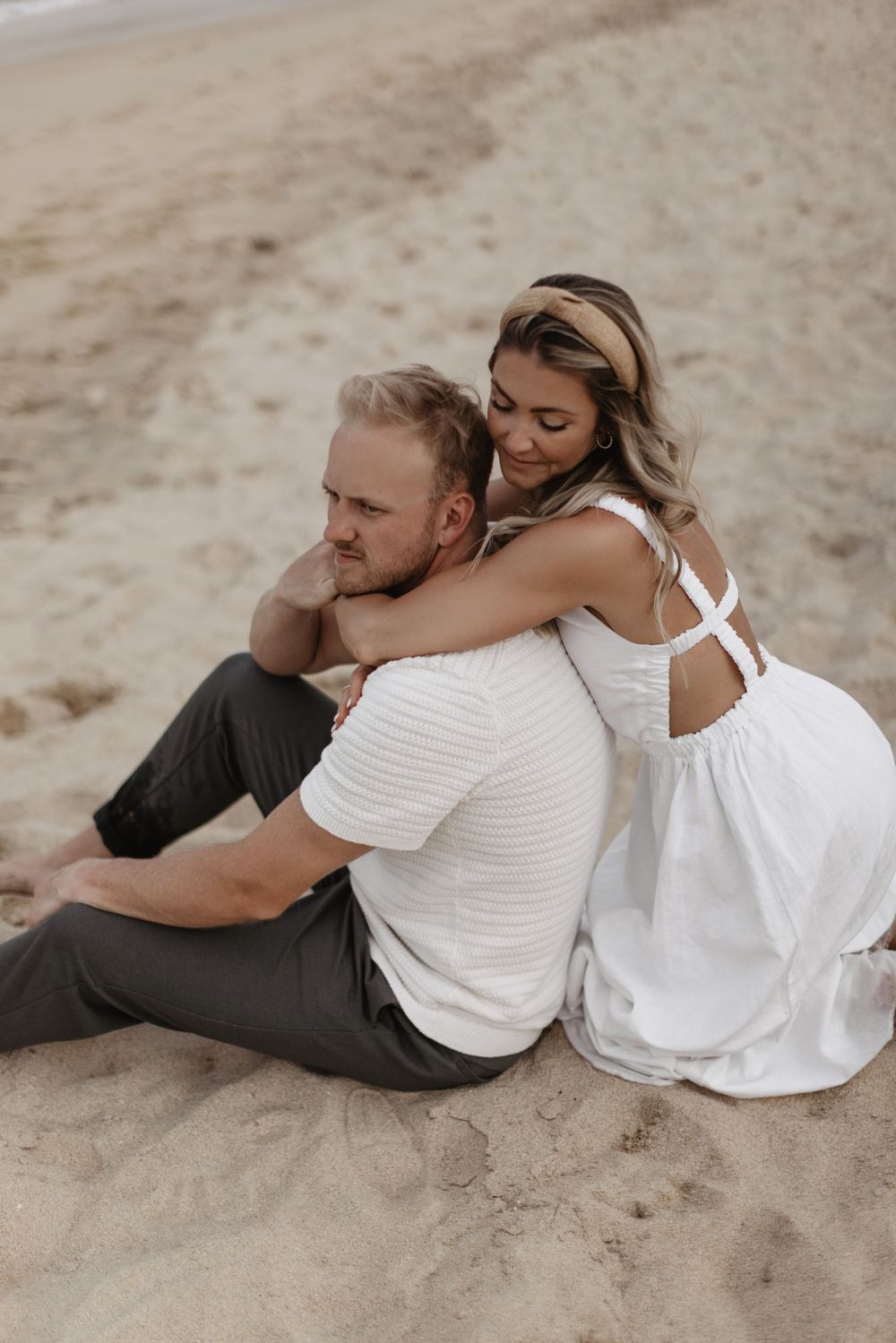 Couple sitting back to back on sandy beach in casual white and black outfits showing intimate romantic moment.