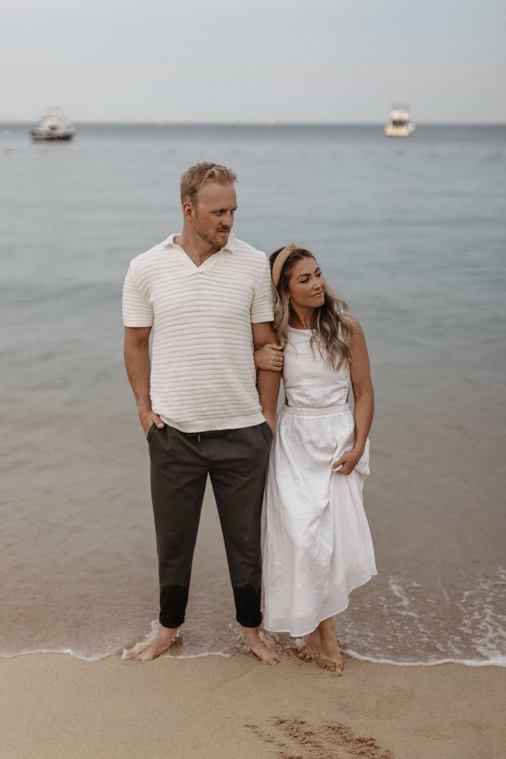 A couple stands together at the water's edge on a sandy beach, dressed in white and beige casual attire.