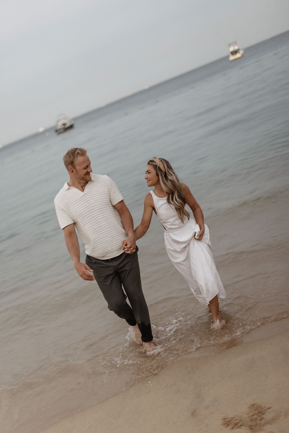 A couple in white and casual attire playfully splashes in the shallow ocean water at sunset.