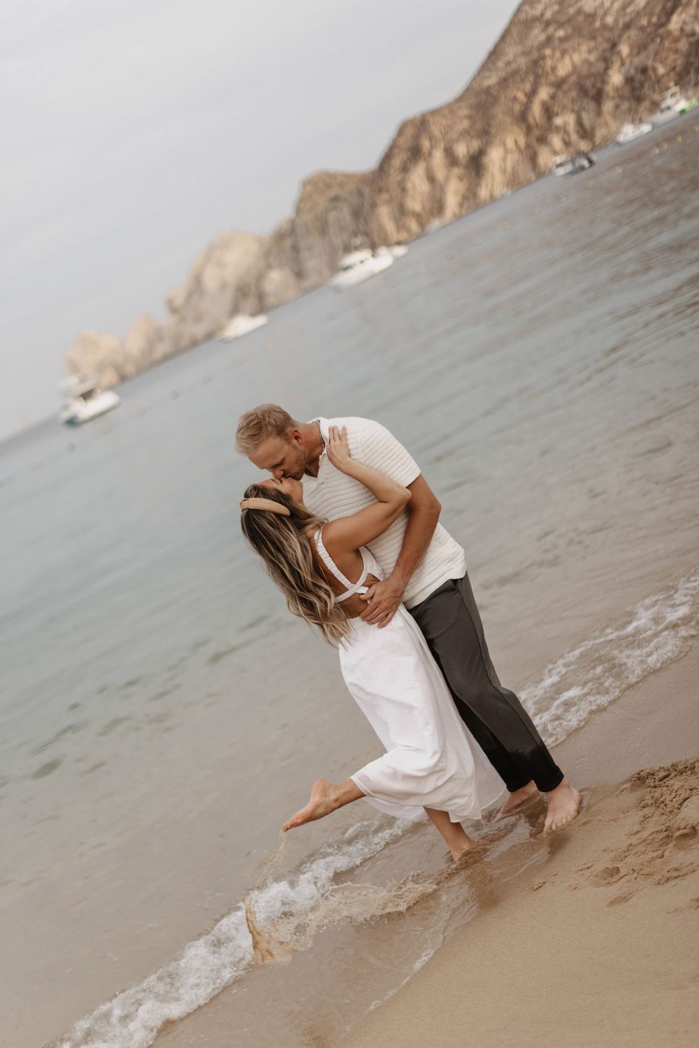 A romantic couple embraces while standing in the ocean waves at the beach with cliffs in the background.