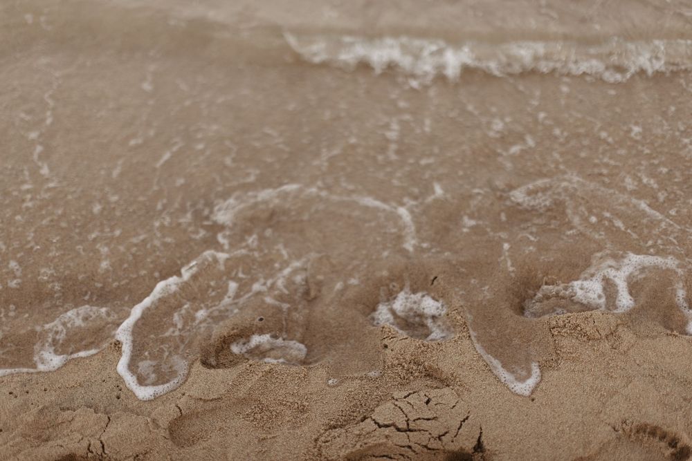 A long panoramic view of brown sandy beach with white seafoam washing up on the shore.