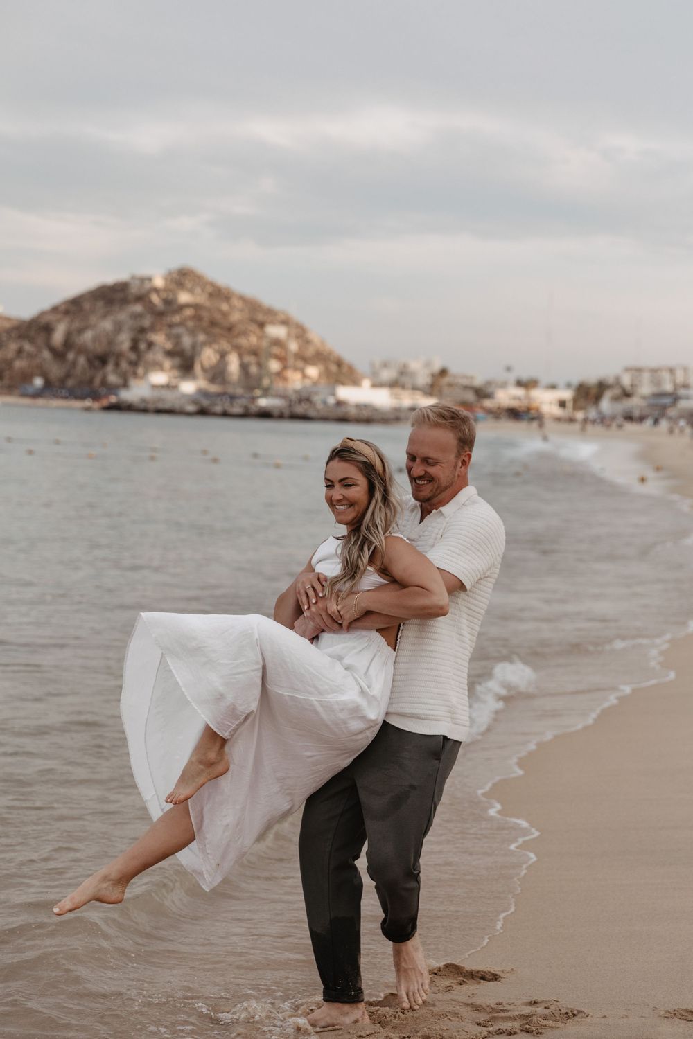 A couple in white and beige attire share a romantic moment on a sandy beach with mountains in the background.
