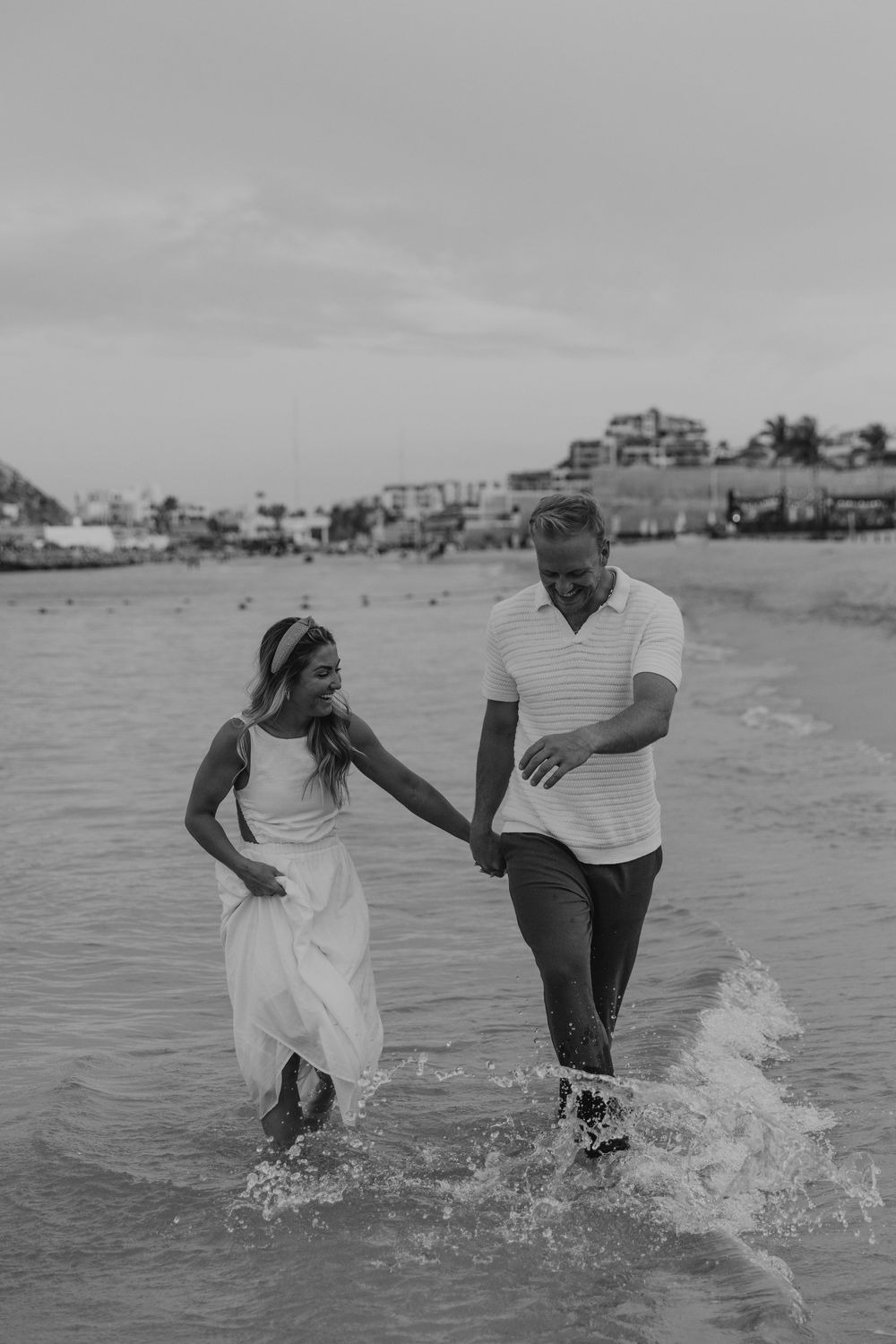 A couple playfully splashes through shallow ocean waves at the beach in black and white photography.
