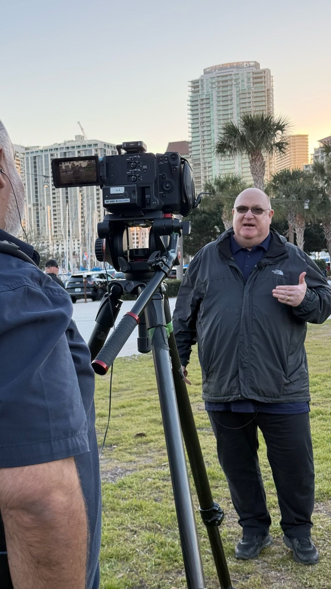 Man being filmed on camera tripod outdoors with city skyline and palm trees in background at dusk.