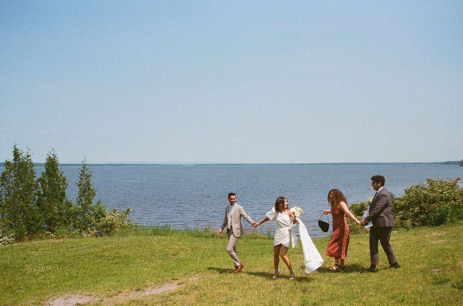 Two couples in formal attire hold hands while walking along a scenic waterfront lawn under a clear blue sky.