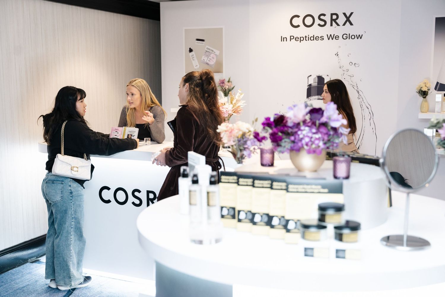 Customers browsing COSRX skincare products at a bright retail display with purple flowers and mirrors.