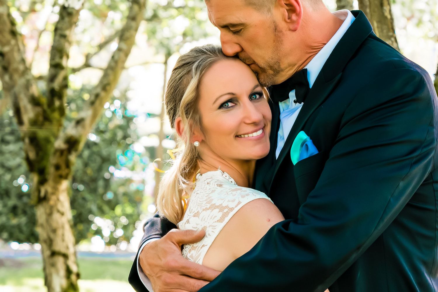A groom in a tuxedo kisses his smiling bride in a white lace dress outdoors among trees on their wedding day.