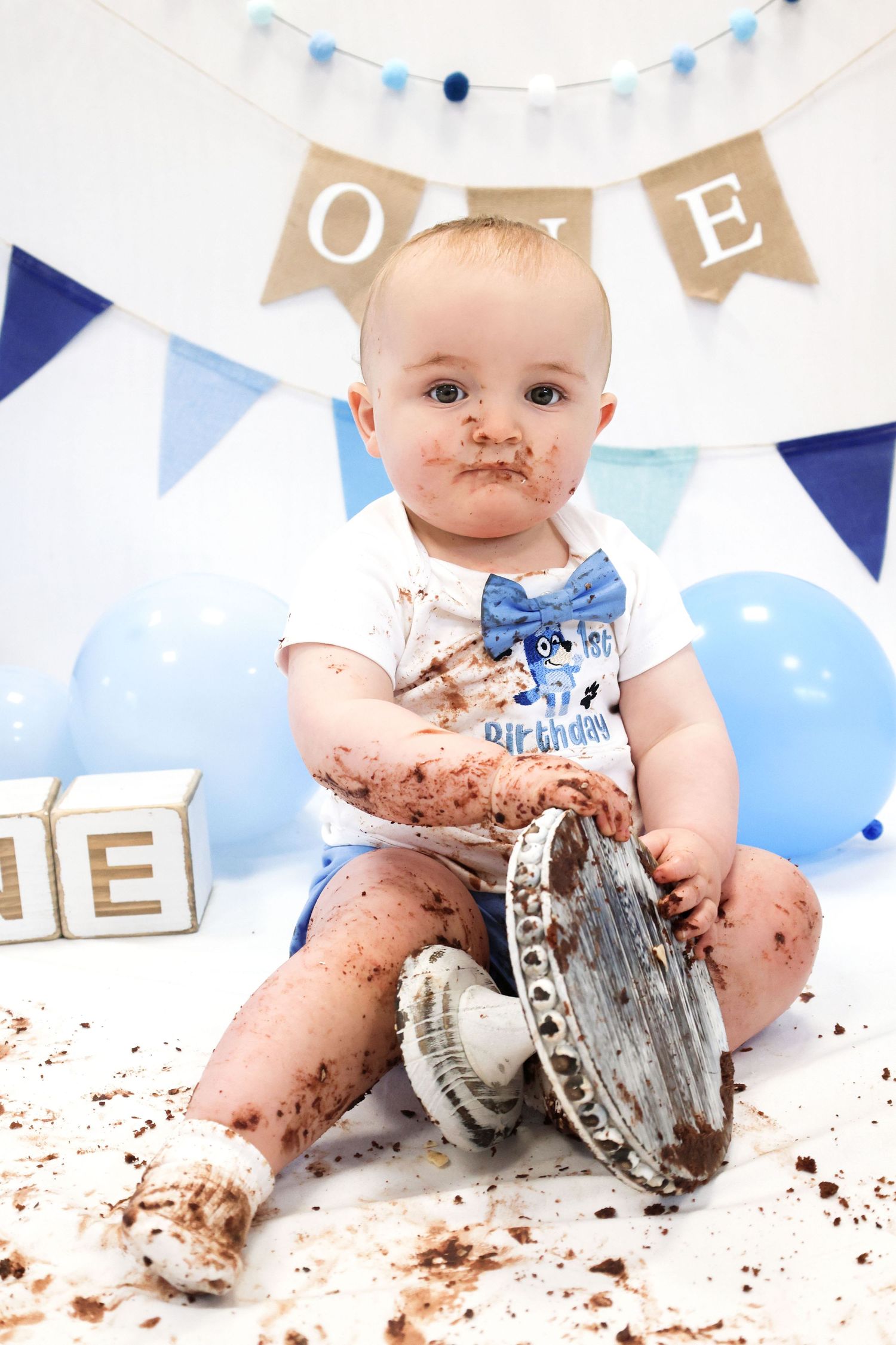 Baby in blue and white decorations smashing cake during first birthday celebration with chocolate smeared everywhere.
