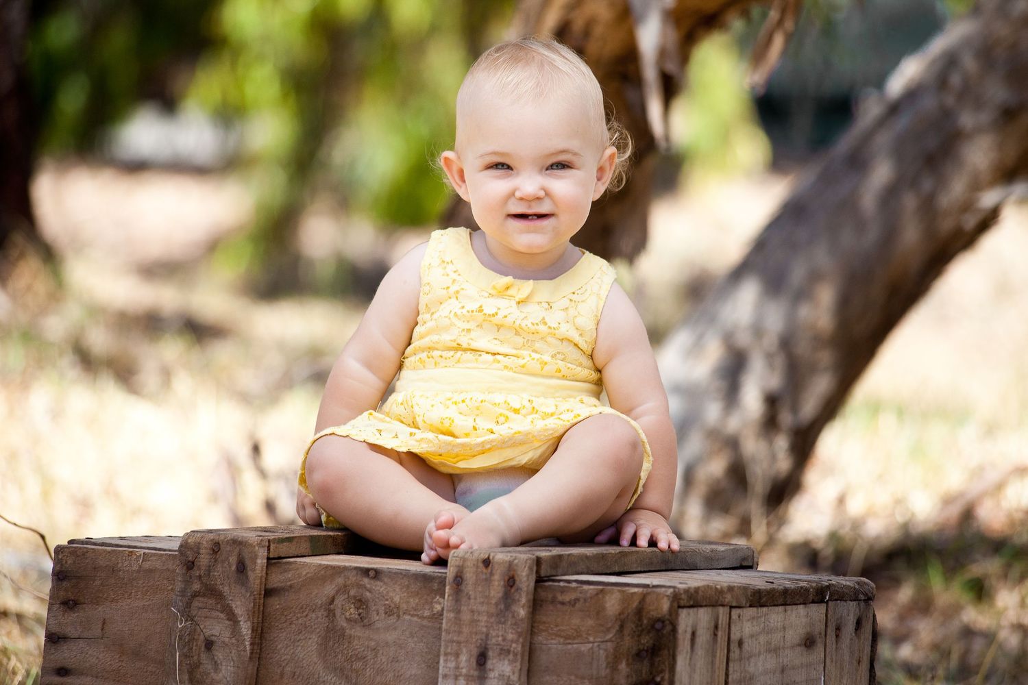 A small child sits outdoors on a rustic wooden bench surrounded by trees and natural scenery.