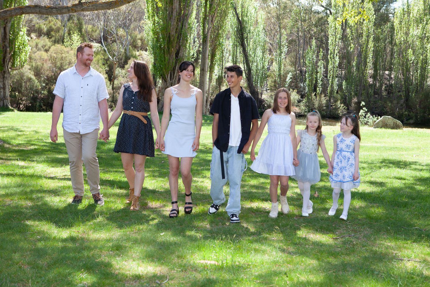 Family group walks together on lush green lawn near willow trees on sunny summer day.