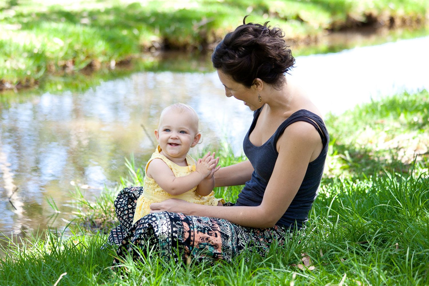 A mother and child sit by tranquil pond edge on grassy bank during outdoor photo session.