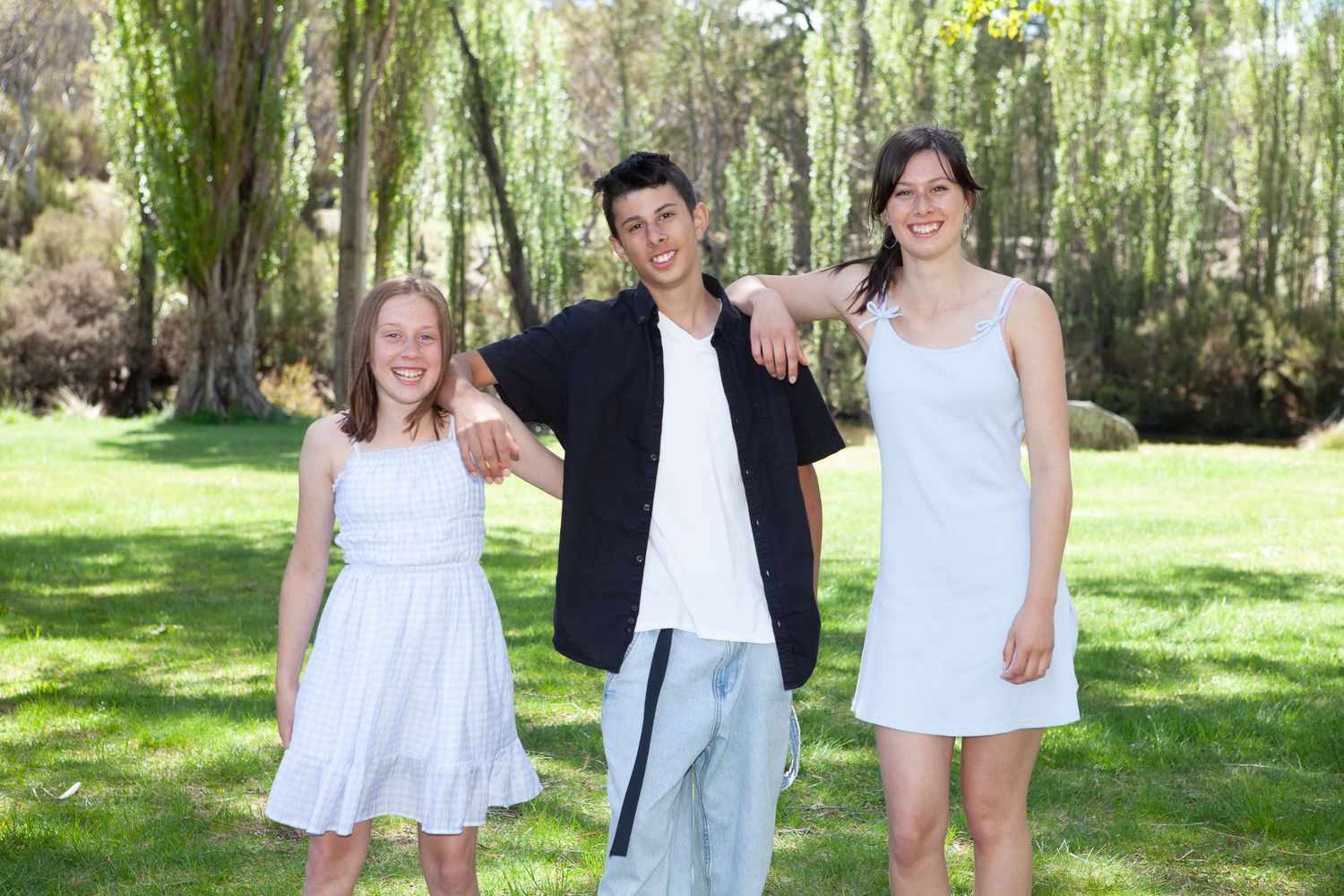 Three siblings in white and black outfits pose together on a sunny day with weeping willow trees in the background.