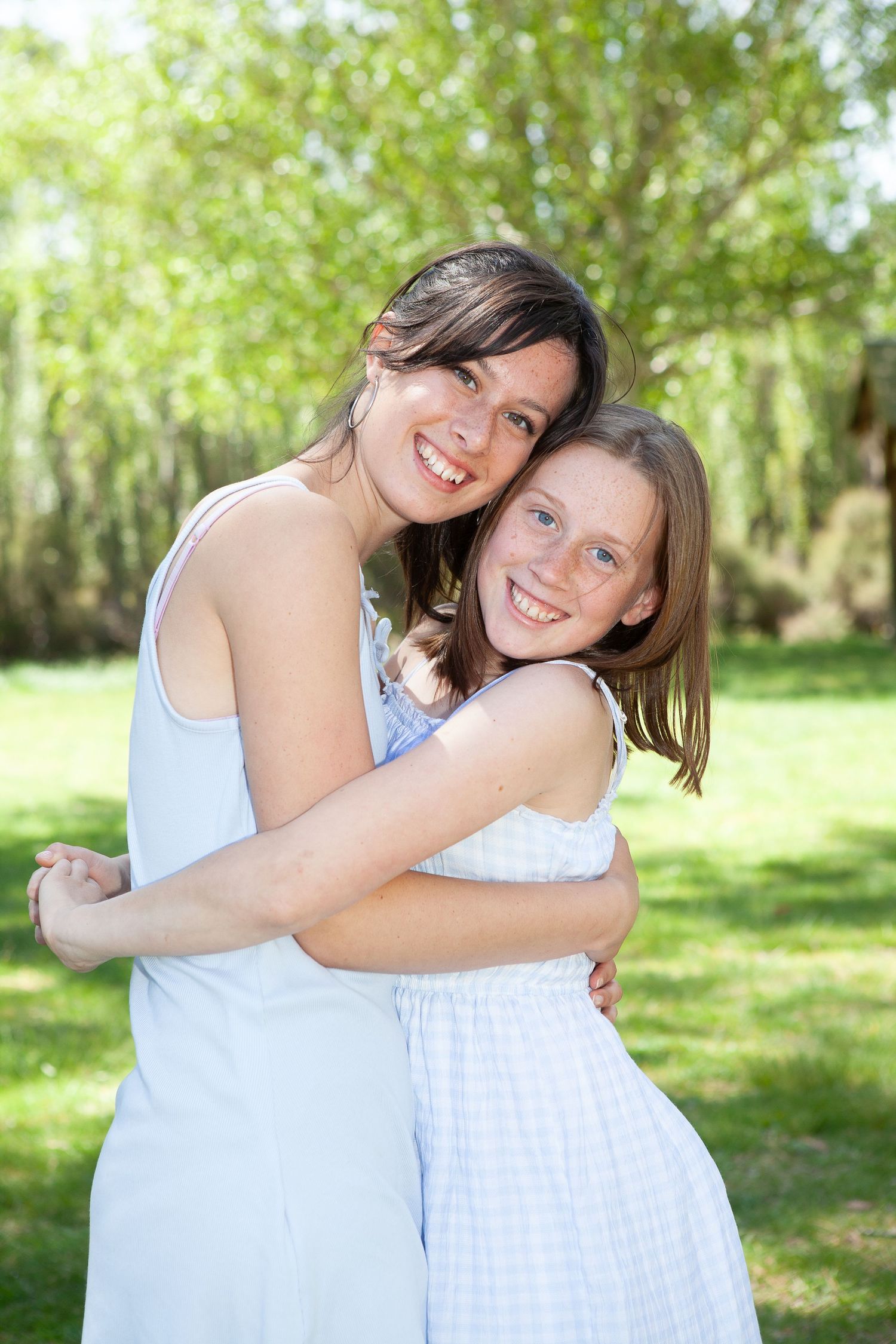 Two people in white dresses embrace and laugh together in a sunny green garden setting.
