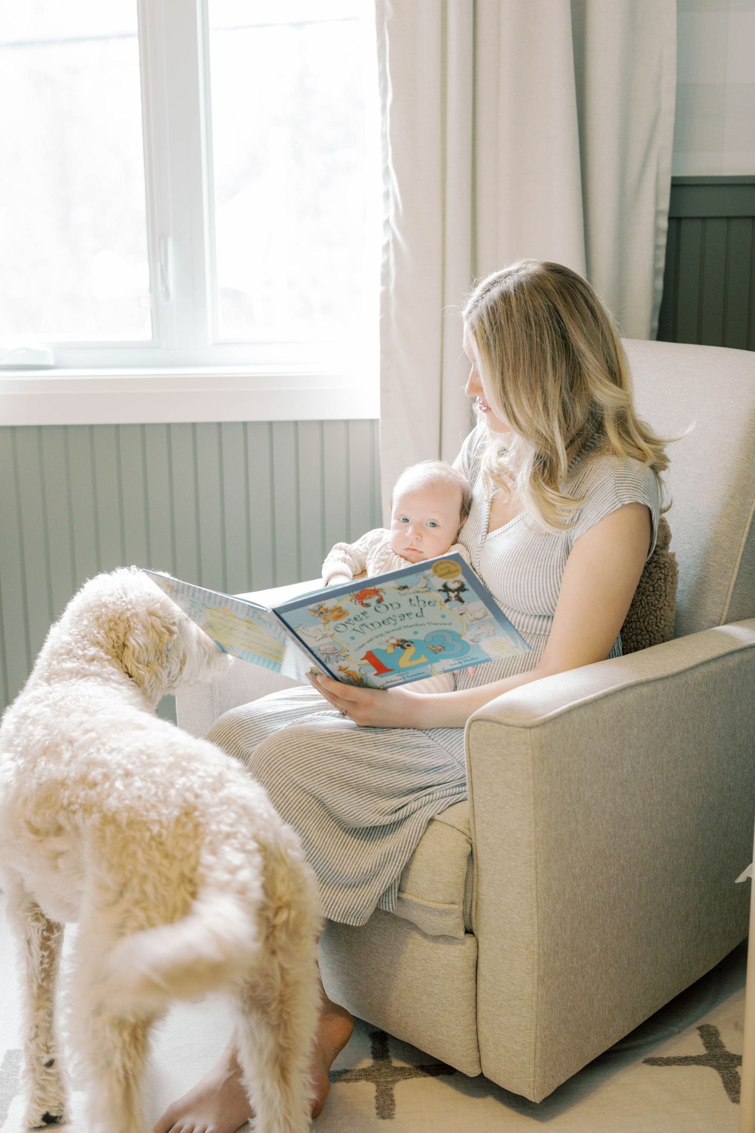 Blonde mother reads colorful picture book to newborn in glider chair as white goldendoodle looks on.