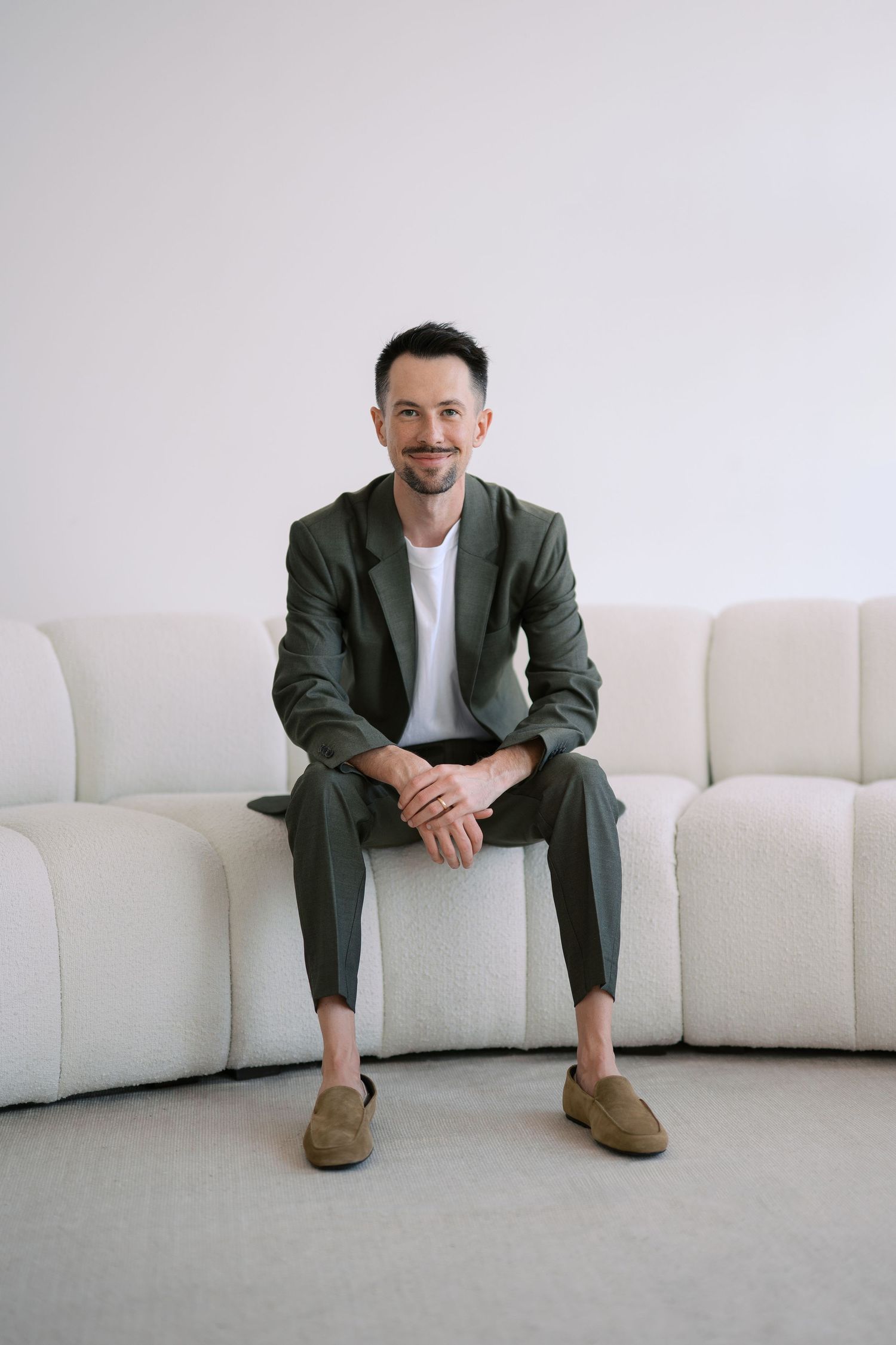 A person in a dark blazer and white shirt sits on a modern white sectional sofa in a minimalist setting.