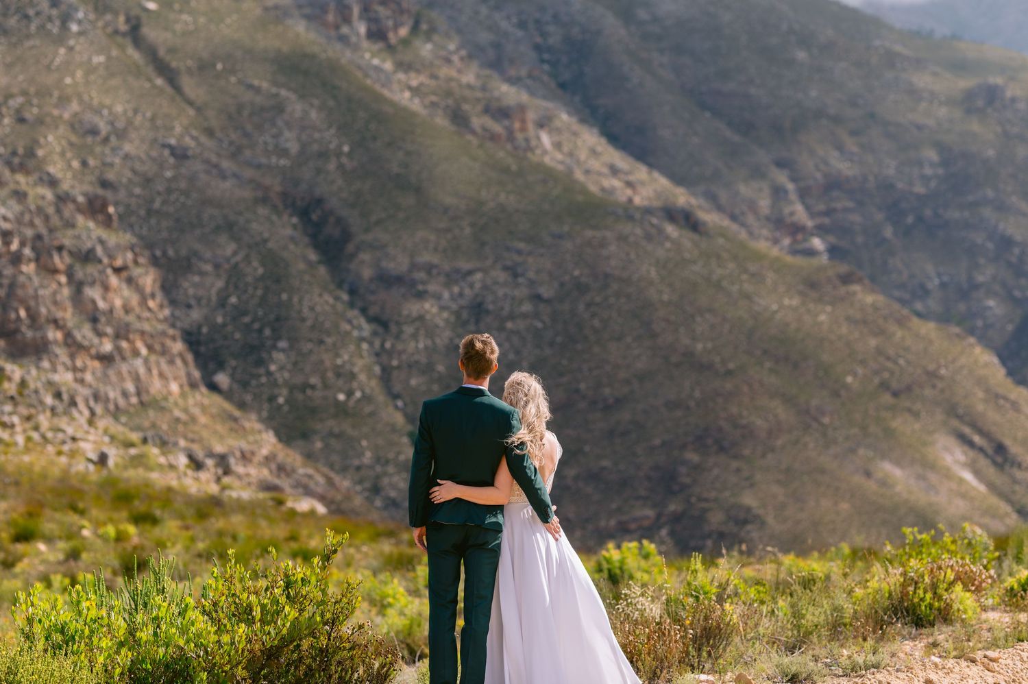 Bride and groom embrace while gazing at scenic mountain landscape during outdoor wedding photoshoot.